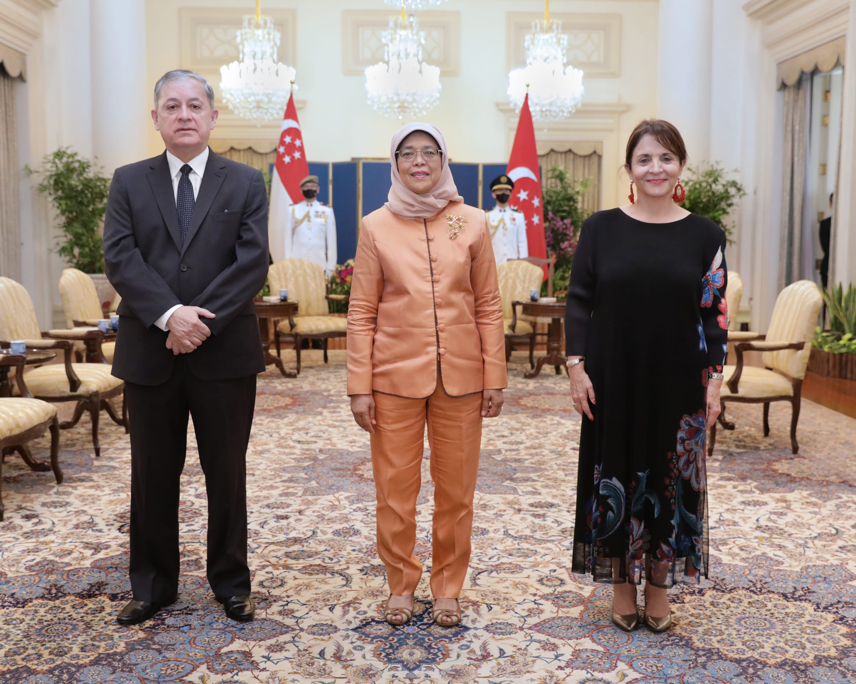Three people stand in front of Singapore flags in a formal room.